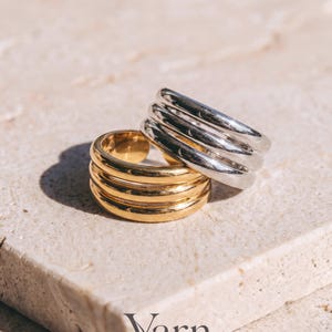 May include: Two multi-band rings, one in a polished gold tone and one in a polished silver tone. Each ring features three smooth, rounded bands connected at the sides to form a single piece. The word Varn is printed in dark gray at the bottom.