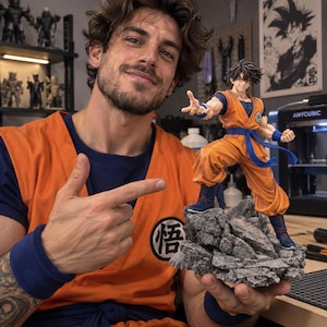 Peut inclure: Une figurine détaillée de style anime représentant un personnage aux cheveux noirs en pointe, vêtu d'un gi d'arts martiaux orange avec une ceinture et des bracelets bleus, posé sur une base rocheuse grise texturée avec un emblème circulaire.