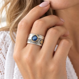 May include: A wide band ring crafted from oxidized silver featuring a teardrop-shaped blue kyanite stone set in a gold bezel. The surface displays a hammered texture with wavy lines and a small cluster of gold-toned metal spheres.