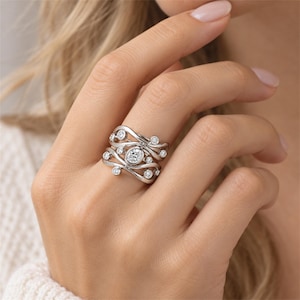 May include: A wide silver-toned ring featuring a multi-layered, wavy band design. The surface is accented with multiple small, round, clear stones and a single, central, oval-shaped clear stone, all set within the flowing metal structure.