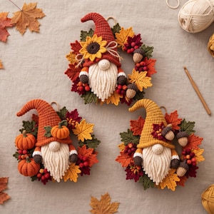 Op de afbeelding: Drie gehaakte kabouter-wandhangers met herfstthema's. Elke kabouter heeft een lange baard, een puntmuts en is omringd door een krans van gehaakte esdoornbladeren, bessen, pompoenen, zonnebloemen en eikels in tinten oranje, geel en rood.