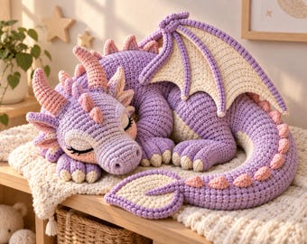 May include: A handcrafted crochet dragon plush toy in shades of lavender and cream. The dragon is depicted in a sleeping position with closed eyes, featuring soft pink spikes along its back and tail, and large, detailed wings with cream-colored webbing.