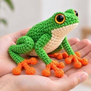 May include: A crocheted frog toy featuring a bright green back, a cream-colored underbelly, and vibrant orange feet with rounded toes. The frog has large, prominent black eyes with orange irises, crafted with detailed yarn stitching throughout.