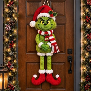 May include: A crocheted green character wall hanging featuring a red Santa hat with white trim, a red and white striped scarf with a small bell, and red curled elf shoes. The figure has a textured green body and is suspended by a string.