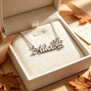 Puede incluir: Un collar de tono plateado con el nombre Alice en letra cursiva, unido a una cadena delicada. El colgante incluye un diseño detallado de hoja de arce al final del nombre. El collar se presenta dentro de una caja blanca abierta.
