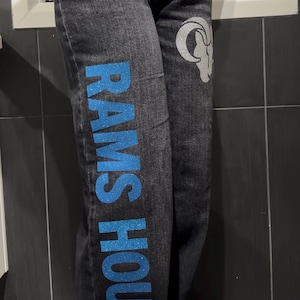 May include: Black denim jeans featuring a vertical blue glitter text graphic that reads RAMS HOUSE on the right leg and a silver glitter ram head logo on the left thigh. The jeans have a standard button fly closure and a classic five-pocket design.