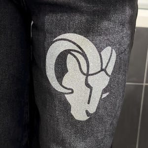 Könnte beinhalten: Schwarze Jeans aus Denim mit einem großen, silbernen, glitzernden Grafikdruck eines Widderkopfes im Oberschenkelbereich. Das Widder-Design ist mit klaren Linien und einem markanten, geschwungenen Horn stilisiert und hebt sich vom Stoff ab.