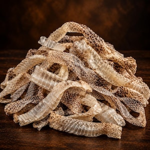 May include: A pile of shed snake skin featuring a translucent, papery texture with a repeating scale pattern. The skin displays natural shades of beige, tan, and dark brown, with elongated, curled strips layered together in a dense, irregular mound.