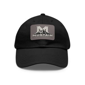 May include: A black baseball cap featuring a rectangular grey patch on the front. The patch displays a white letter M above a mountain range graphic, with the text MUSTAIN and the phrase BUILT FOR MORE printed in white below the mountain.