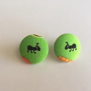 May include: Two round, lime green fabric buttons. Each button features a black ant illustration. The buttons have a textured appearance and orange and yellow accents. These buttons are suitable for sewing or crafting projects.