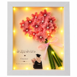 Könnte beinhalten: Ein weißer Schattenfugenrahmen mit einem Strauß aus rosa Pfeifenreiniger-Blumen mit Perlen in der Mitte, gebunden mit einem rosa Band. Darunter eine Illustration eines Absolventen in Talar mit Blumen sowie der Text "Chaymae", "MAVO GRADUATE 2026" und arabische Schriftzeichen. Das Display ist mit warmen LED-Lichtern beleuchtet.