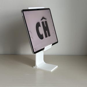 Può includere: Un supporto bianco per tablet che sostiene un tablet argentato con uno stilo nero fissato sul bordo superiore. Lo schermo del tablet mostra uno sfondo grigio chiaro con il testo nero CH con un accento circonflesso sulla lettera C al centro.