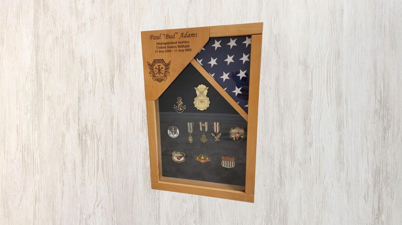 May include: A wooden shadow box with a folded American flag and a display of military medals and insignia. The box is engraved with the name "Paul "Bud" Adams" and the text "Distinguished Service, United States Military, 10 July 1968 - 01 May 1992".
