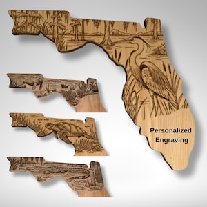 May include: Wooden wall art shaped like Florida, featuring engraved wildlife scenes. The largest piece shows a heron and the text "Personalized Engraving." Smaller pieces depict other Florida animals. The wood has a natural, light brown colour.