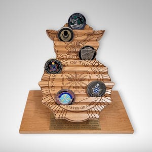 Puede incluir: Soporte de exhibición de madera con forma del emblema de la Fuerza Aérea de los Estados Unidos, con ranuras para guardar monedas de desafío. El soporte está hecho de madera marrón claro y presenta las palabras "Department of the Air Force" y "United States of America".