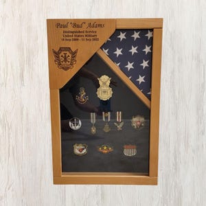 May include: A wooden shadow box with a framed American flag and a black background. The shadow box has a plaque with the text "Paul "Bud" Adams Distinguished Service United States Military 18 Sep 2008 - 13 July 2022" and a collection of military medals and pins.