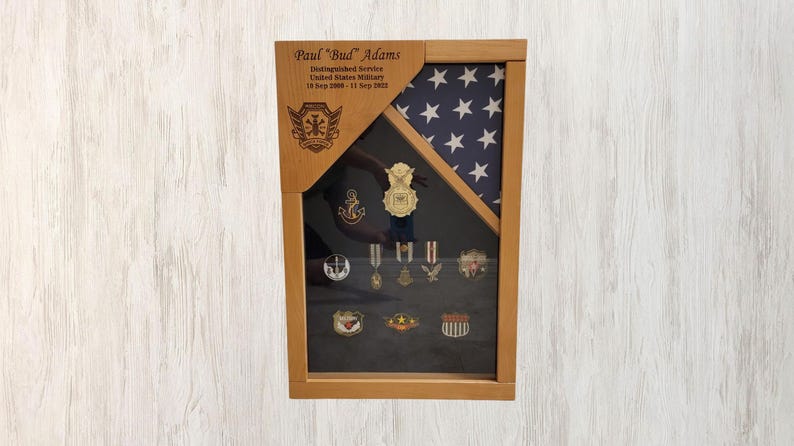 May include: A wooden shadow box with a framed American flag and a black background. The shadow box has a plaque with the text "Paul "Bud" Adams Distinguished Service United States Military 18 Sep 2008 - 13 July 2022" and a collection of military medals and pins.