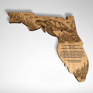 May include: Wooden plaque in the shape of Florida, featuring an engraved sea turtle and aquatic plants. The plaque includes engraved text. The design is a light brown color.