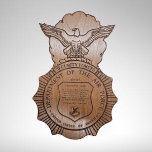 Puede incluir: Una placa de madera con un diseño grabado con láser del emblema de las Fuerzas de Seguridad de la Fuerza Aérea de los Estados Unidos. La placa presenta un escudo con las palabras "Department of the Air Force" y "United States of America" que lo rodean. El escudo también incluye un emblema más pequeño de un halcón dentro de un círculo. La placa incluye el texto "Air S/Sgt.  A/1C Michael Thomas" y un breve mensaje de agradecimiento.