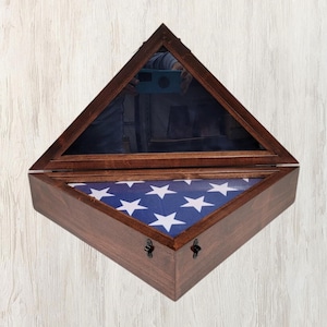 Może przedstawiać: Drewniana, trójkątna skrzynka z szklanym wierzchem, prezentująca złożoną flagę amerykańską z białymi gwiazdami na niebieskim polu. Skrzynka ma dwa zawiasy i zatrzask.