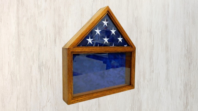 May include: A wooden flag display case with a glass front and a folded American flag inside. The case is shaped like a house and has a dark wood finish.