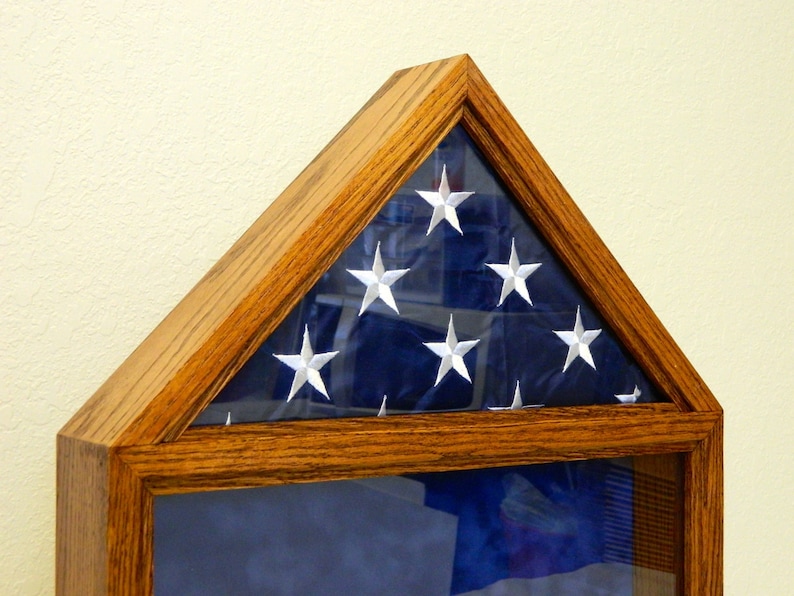 May include: A wooden flag display case with a glass front. The case is in the shape of a triangle and has a dark blue flag with white stars inside. The case is made of oak wood and has a natural finish.