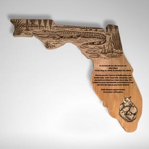 Op de afbeelding: Houten plaquette in de vorm van Florida, met een gegraveerde alligatorscène en tekst. De plaquette bevat een bedankbericht en een tekening van een gespierde neushoorn. Het hout heeft een natuurlijke, lichtbruine kleur.