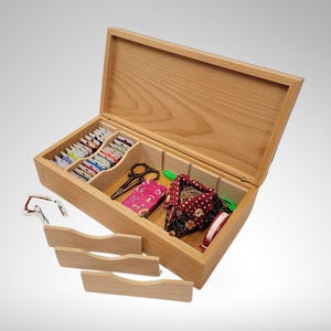 May include: A wooden box with a hinged lid, containing a variety of sewing supplies, including spools of thread, scissors, and a pincushion. The box has two small drawers on the front.