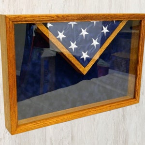 May include: A wooden shadow box with a glass front, displaying a folded American flag. The flag is folded in a triangular shape and is centered in the box. The box is a light brown color and has a simple design.