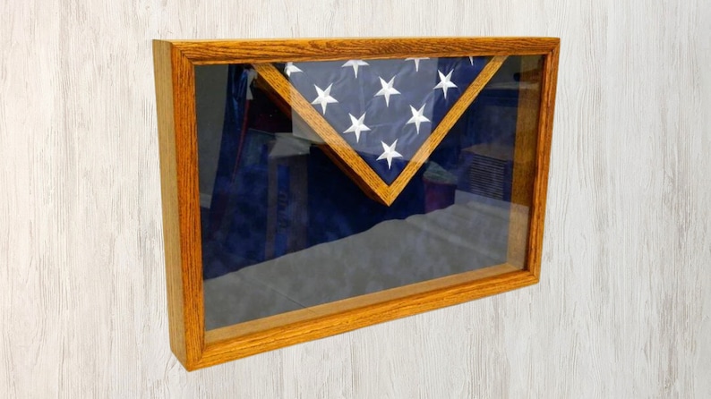 May include: A wooden shadow box with a glass front, displaying a folded American flag. The flag is folded in a triangular shape and is centered in the box. The box is a light brown color and has a simple design.