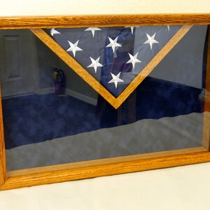 May include: A wooden flag display case with a glass front. The case is stained a dark wood color and has a blue velvet backing. A folded American flag is displayed inside the case.
