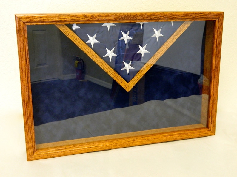 May include: A wooden flag display case with a glass front. The case is stained a dark wood color and has a blue velvet backing. A folded American flag is displayed inside the case.