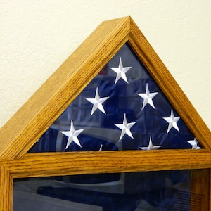 May include: A wooden shadow box with a glass front, displaying a folded American flag. The flag is blue with white stars and is folded in a triangular shape.