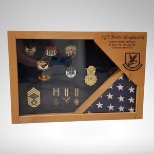 May include: A wooden shadow box with a folded American flag and a display of military medals and patches. The box is engraved with the text "SGT Marie Stingaworth United States Military 01 May 98 - 29 Mar 03 "Defend Our Way" "