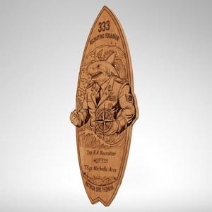Könnte beinhalten: Holzplakette in Surfbrettform mit einem geschnitzten Design. Das Design zeigt einen Hai in Militäruniform, der einen Kompass hält, mit dem Text "333 Recruiting Squadron" und "Top EA Recruiter".