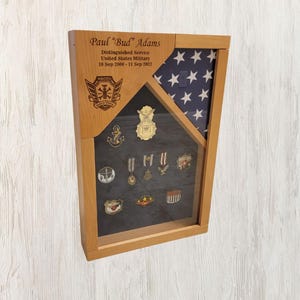 May include: A wooden shadow box with a United States flag, a personalized inscription, and a display of military medals and pins. The inscription reads "Paul 'Bud' Adams Distinguished Service United States Military 18 Sept 1998 - 11 Sept 2023".