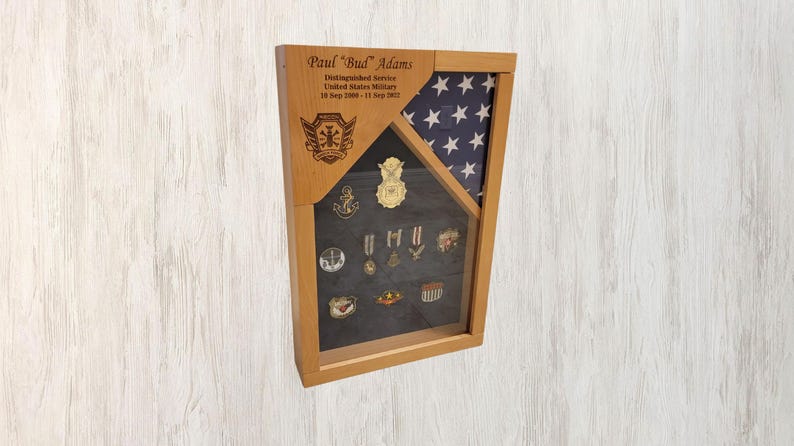 May include: A wooden shadow box with a United States flag, a personalized inscription, and a display of military medals and pins. The inscription reads "Paul 'Bud' Adams Distinguished Service United States Military 18 Sept 1998 - 11 Sept 2023".
