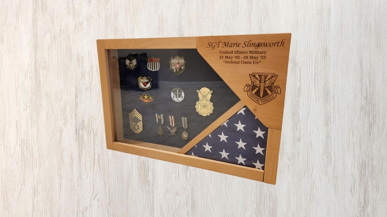 May include: A wooden shadow box with a glass front displaying military medals and a folded American flag. The box is engraved with the text "SGT Marie Slingsworth, United States Military, 23 May 03-20 May 23, "Defend Onto Us."