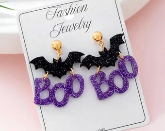 May include: A pair of dangle earrings on a white display card labeled Fashion Jewelry and Vellora. The earrings feature a gold-tone stud, a black glitter bat silhouette, and a purple glitter cutout of the word Boo hanging from the bat.