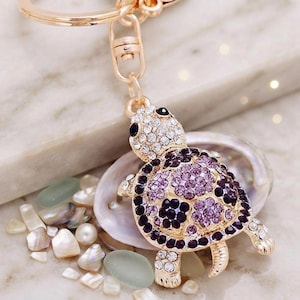 May include: A gold-toned metal keychain featuring a turtle charm encrusted with clear, light purple, and dark purple rhinestones. The turtle has a detailed shell pattern and is attached to a circular metal ring by a small lobster clasp.