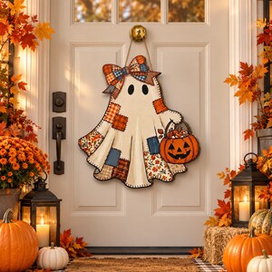 May include: A decorative wooden ghost door hanger featuring a cream-coloured body with various colourful plaid and floral fabric-style patches, a large matching bow at the top, and a small orange jack-o'-lantern bucket held in one hand.