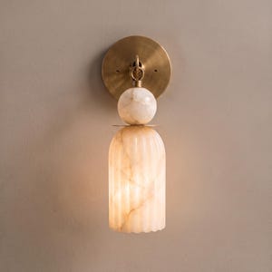 May include: A wall sconce featuring a circular brushed brass backplate, a spherical stone accent, and a cylindrical, vertically fluted translucent stone shade. The light fixture displays warm beige and cream veining throughout the stone components.