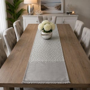 May include: A rectangular table runner featuring a central panel with a repeating white and grey honeycomb pattern, flanked by solid grey end sections with fringe detailing. The runner is positioned lengthwise on a wooden dining table.