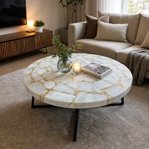 May include: A round coffee table featuring a white stone top with a cracked, mosaic-like pattern filled with gold-toned veins. The table is supported by a black metal frame with thin, angled legs. A glass vase with greenery, a candle, and a book are placed on top.