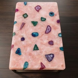 Könnte beinhalten: Ein rechteckiger Couchtisch mit einer glatten, polierten hellrosa Oberfläche, in die verschiedene, unregelmäßig geformte, edelsteinartige Akzente in Blau-, Grün-, Violett- und Magentatönen eingelassen sind, auf einem dunklen Metallrahmen.