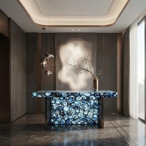 May include: A rectangular console table featuring a surface and base constructed from polished blue agate slices with visible concentric rings. A tall, slender metallic vase containing a bare, dark-colored branch sits on the table surface.