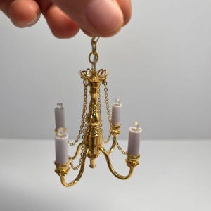 Könnte beinhalten: Ein goldfarbener Miniatur-Kronleuchter mit drei geschwungenen Armen, die jeweils eine kleine weiße kerzenförmige Leuchte tragen. Die Struktur ist an feinen Goldketten befestigt, die mit einem zentralen Stab und einer Öse verbunden sind.