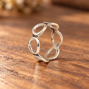 May include: A silver-toned metal ring featuring a series of interconnected circular open loops forming the band. The metal has a polished finish with darkened recessed areas between the circular links, creating a distinct contrast in the design.