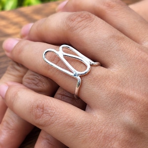 Può includere: Un anello in argento caratterizzato da un design a filo aperto, allungato e curvo che si estende verticalmente lungo il dito. La fascia in metallo lucido forma una figura fluida e continua con bordi arrotondati, creando una sagoma vuota.