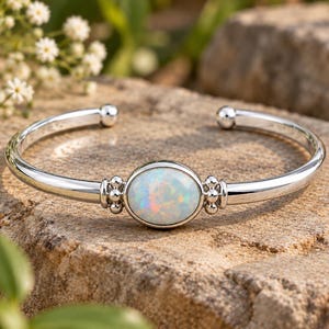 Pode incluir: Uma pulseira de punho em prata com uma pedra de opala oval e iridescente ao centro. A banda de metal polido termina em pequenas esferas e o engaste é decorado com minúsculas contas de prata em ambos os lados da opala.